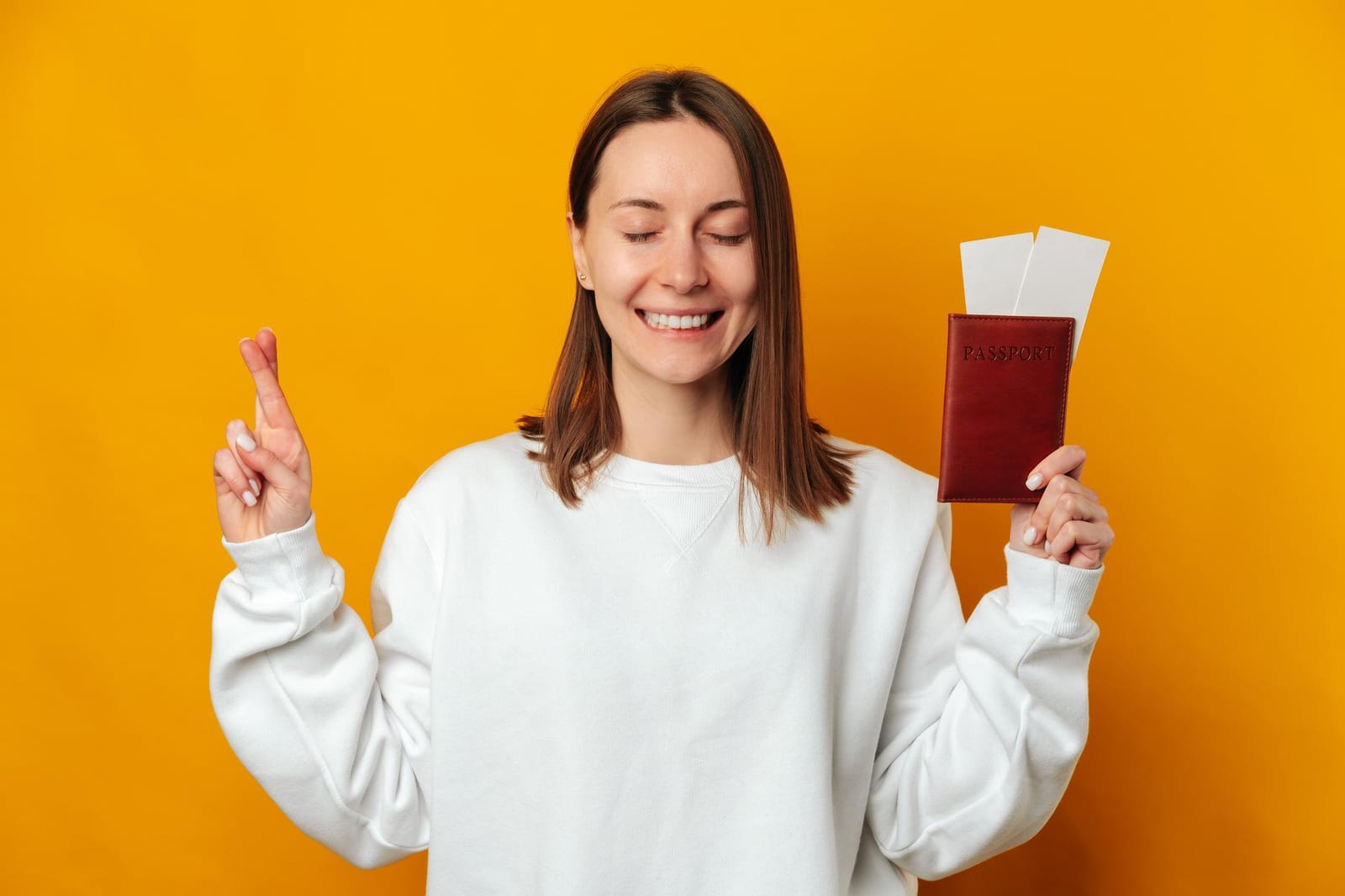 Mulher sobre fundo amarelo cruza os dedos enquanto segura uma carteira de passaporte com passagens.