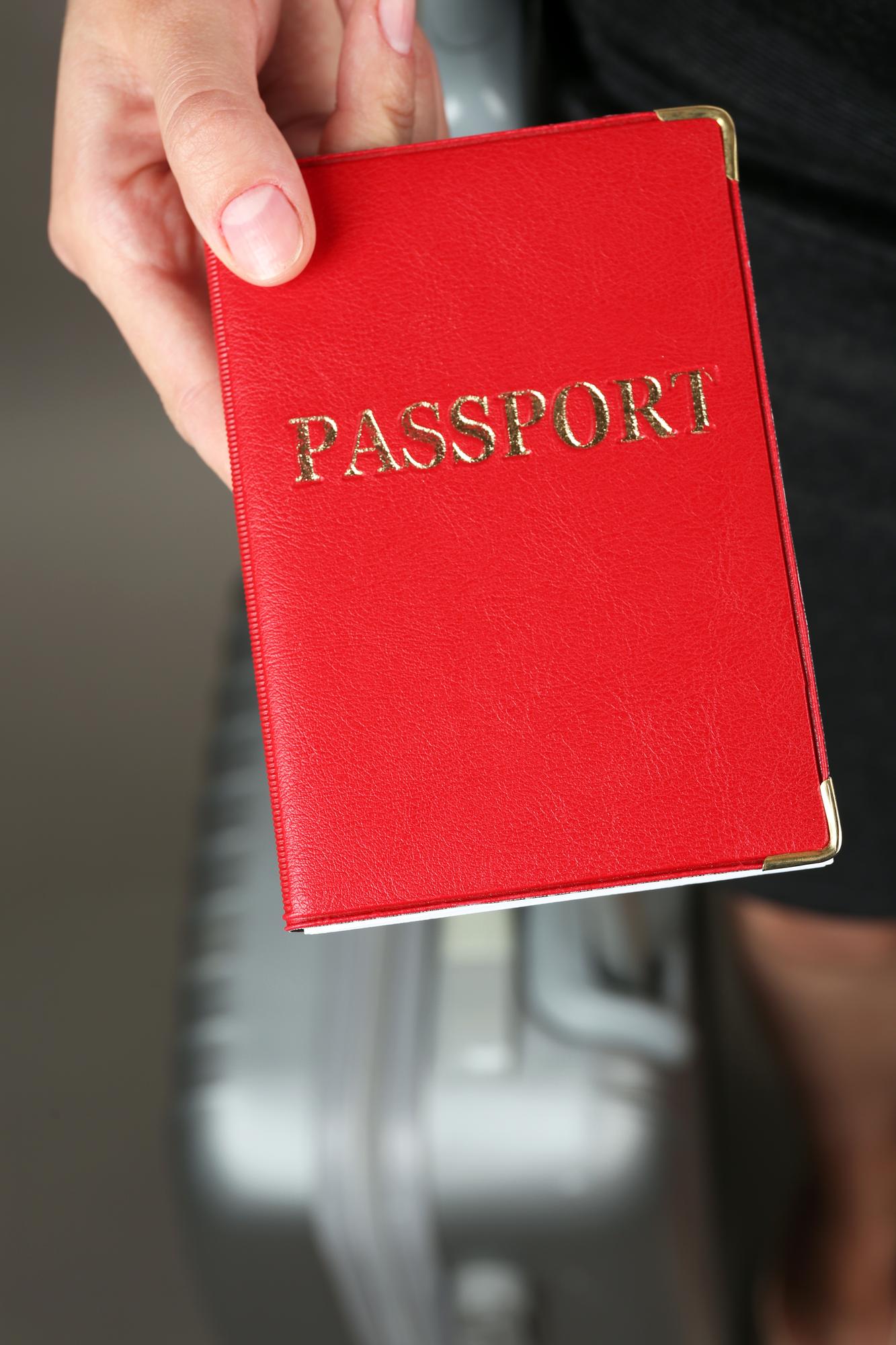 Mão segura a capa vermelha de um passaporte à frente de uma mala de viagem.